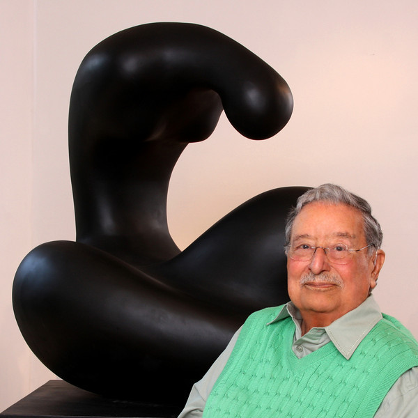 Portait de Manuel Carbonell assis à côté d'une de ses oeuvres de couleur noire.