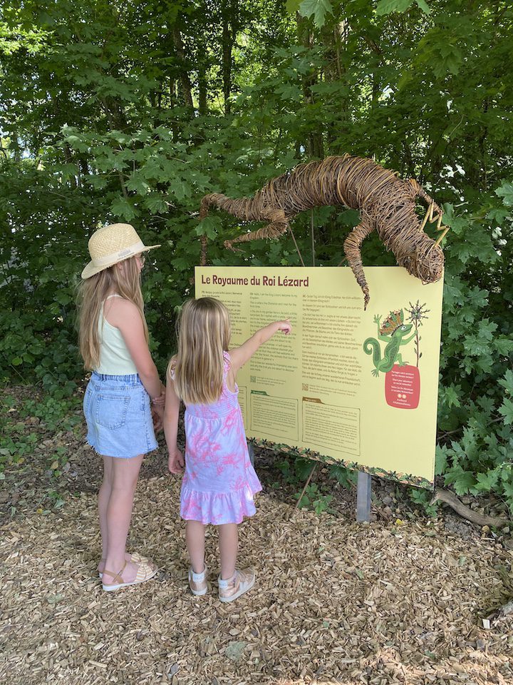 2 fillettes lisant le panneau d'accueil du Royaume du Roi Lézard du Château de Vullierens en été.